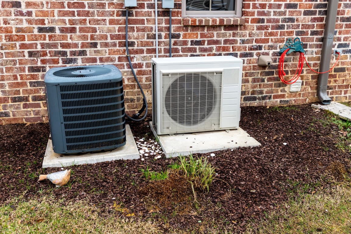 Condenser and a ductless heat pump outdoor unit on pads beside a brick wall