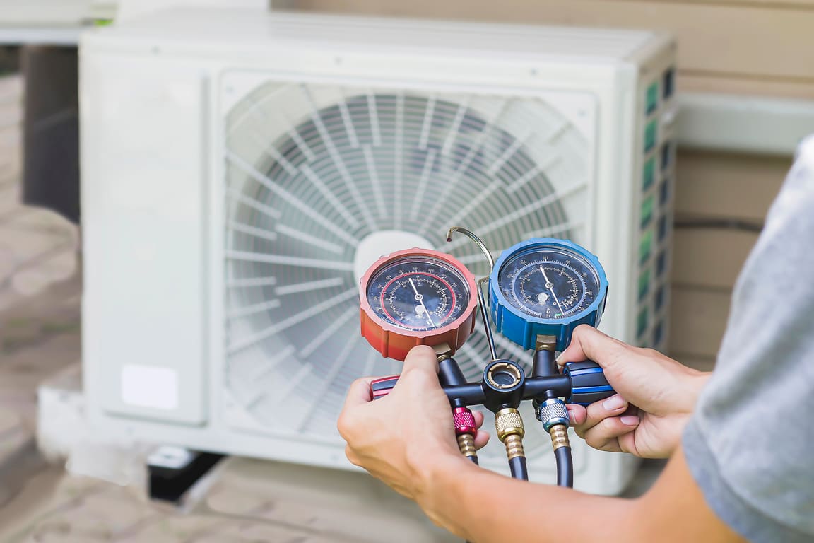 Hands holding a refrigerant gauge set in front of an outdoor heat pump unit